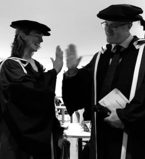 Two people in graduation gowns and caps are giving a high-five. One holds a camera and papers. They seem to be celebrating.